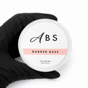 A hand wearing a black glove holds a round white container of ABS Rubber Base TPO and hema free against a plain white background.