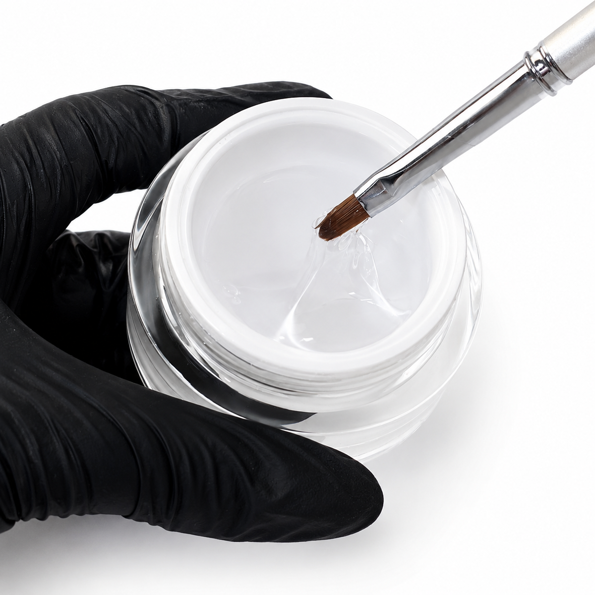 A hand wearing a black glove holds an open clear jar of glossy white gel with a metal brush dipping into the substance against a white background.